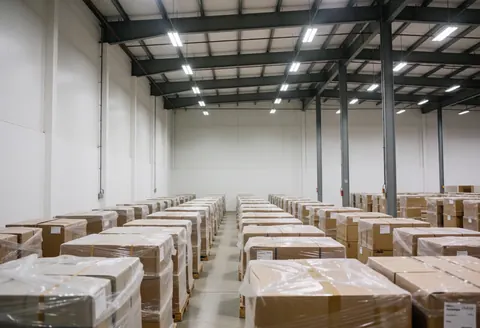 Vue d'un entrepôt de meubles moderne et bien rangé, avec des caisses empilées sur des palettes, un éclairage industriel et des murs blancs.