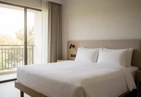 Chambre d'hôtel minimaliste avec un grand lit, des rideaux et une vue sur le balcon avec des arbres. Ambiance calme et lumineuse.
