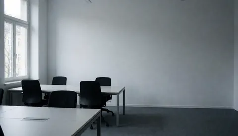Espace de bureau moderne et vide, équipé de tables et de chaises noires, murs gris et lumière naturelle entrant par une fenêtre, servant de fond pour du texte.