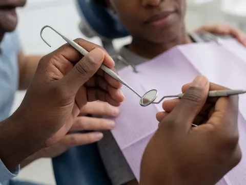 Mains d'un professionnel de la santé tenant des outils dentaires, avec un patient en arrière-plan dans un contexte clinique.
