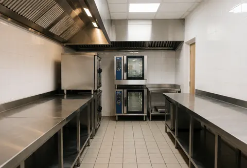 Cuisine professionnelle vide avec comptoirs en acier inoxydable, murs et sol carrelés, casseroles et fours modernes, éclairage lumineux et propre.
