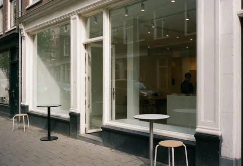 Façade d'agence de marketing à Amsterdam avec grandes baies vitrées, tables et chaises en extérieur, ambiance moderne et minimaliste.