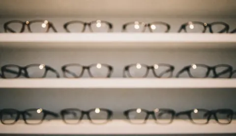 Lunettes à monture sombre posées sur des étagères blanches, avec un fond flou et des lumières chaudes, idéal pour ajouter du texte commercial.