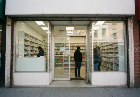 Façade de pharmacie avec clients entrant et vitrine de produits, ambiance urbaine et propre.
