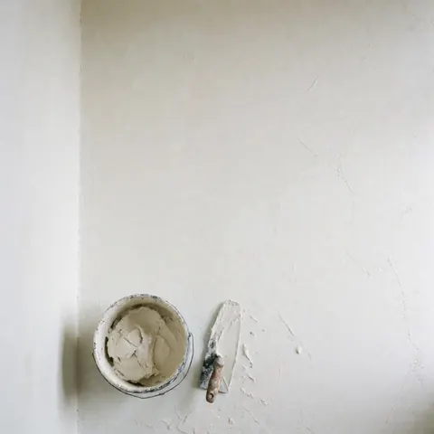 Mur blanc fraîchement enduit accompagné d'un seau d'enduit et d'un rouleau, éclairage naturel doux, idéal pour la décoration ou la construction.