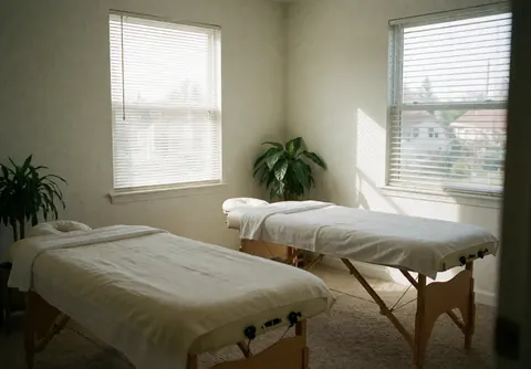 Salle de massage propre et calme avec deux lits couverts de draps blancs, plantes vertes et lumière naturelle par des fenêtres avec stores.