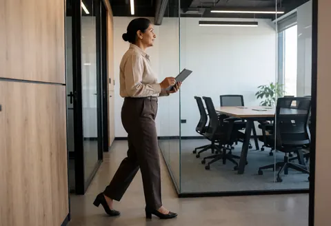 Une femme d'affaires avance dans un couloir de bureau en tenant une tablette, regardant vers une salle de réunion équipée de tables et de chaises.