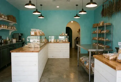 Vue intérieure d'une boulangerie avec des murs turquoise, des comptoirs blancs, des produits en vitrine et des étagères métalliques contenant du pain et des pâtisseries.