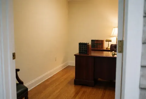 Vue depuis la porte d'un bureau d'avocat vide comprenant un bureau en bois, des livres, une lampe, des murs crème et un sol en bois poli.