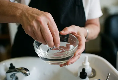 Des mains en train de mélanger un liquide dans un bol en verre posé sur un lavabo dans un salon de manucure, avec un fond flou et une lumière naturelle.