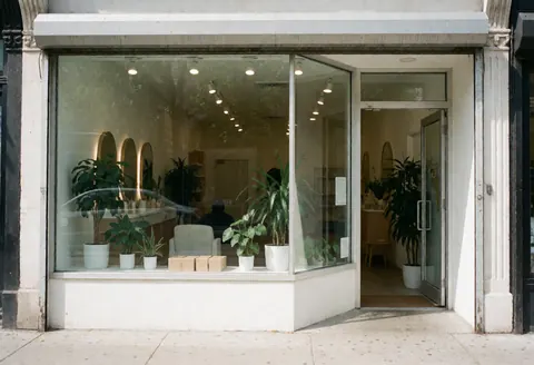 Façade de salon de coiffure avec grandes baies vitrées, plantes vertes et porte d'entrée. Intérieur lumineux et accueillant pour les clients.