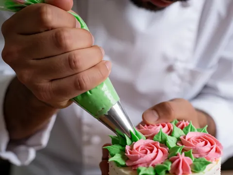 Un petit gâteau orné de fleurs en glaçage rose et de feuilles vertes, en cours de décoration par un chef utilisant une poche à douille.