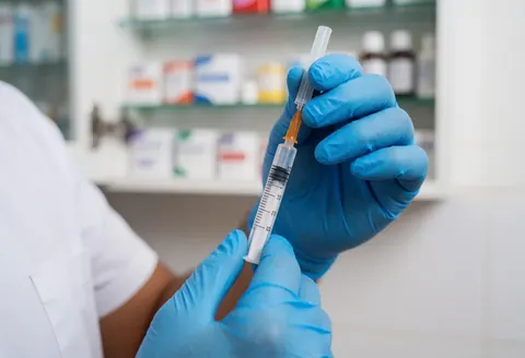Une main portant un gant bleu tient une seringue remplie de vaccin sur fond flou d'étagères de médicaments.