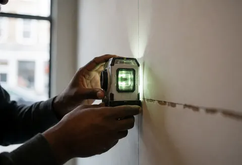 Main d'un travailleur utilisant un niveau laser pour tracer une ligne droite sur un mur blanc dans un contexte de construction intérieure avec éclairage professionnel.