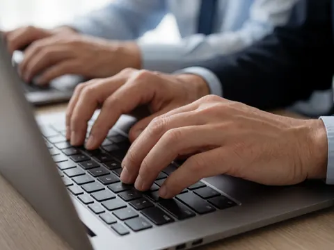 Profilé en chemise et cravate, des mains saisissent les touches d'un ordinateur portable dans un bureau moderne.