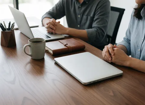 Deux personnes travaillant à une table en bois avec un ordinateur portable, une tasse de café, un cahier et un porte-stylos éclairés par la lumière naturelle.