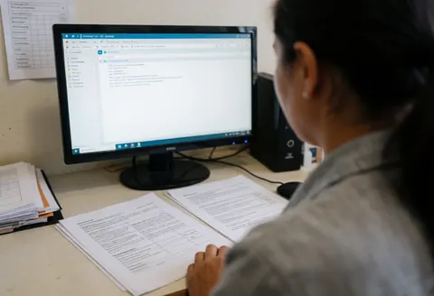 Vue arrière d'une femme concentrée devant son écran d'ordinateur, des papiers posés sur le bureau dans un cadre professionnel réaliste.