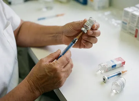 Gros plan sur des mains tenant une seringue et un flacon de médicament posés sur une surface blanche propre, évoquant un environnement clinique professionnel.