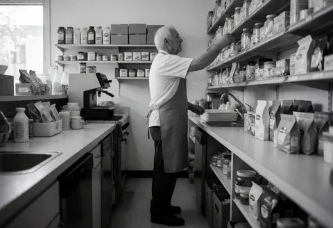 Un homme âgé portant un tablier atteint un produit sur une étagère dans un magasin, avec des produits visibles en arrière-plan.