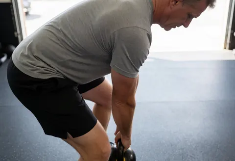 Un homme d'âge mûr effectue un exercice de force avec un kettlebell dans un gymnase, vêtu d'un t-shirt gris et de shorts noirs, sous un éclairage naturel.