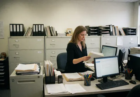 Une femme d'affaires consulte des papiers sur son bureau entouré de classeurs et d'ordinateurs dans un cadre professionnel.