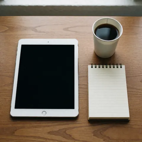 Une tablette, une tasse de café et un cahier posés sur une table en bois dans un espace de travail.