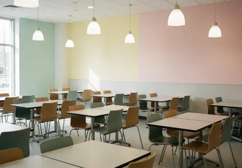 Cafétéria universitaire moderne avec des tables et chaises vides, murs pastel et luminaires suspendus, ambiance propre et accueillante pour les étudiants.
