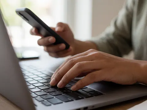 Mains sur le clavier d'un ordinateur portable avec un téléphone à la main, dans un bureau lumineux, ambiance professionnelle.