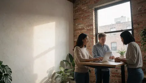 Trois individus collaborant autour d'une table avec un ordinateur portable, dans un espace moderne avec mur de briques et plantes vertes.