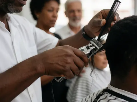 Un barbier coupe les cheveux d'un client à l'aide de ciseaux, dans un salon de barbier avec d'autres clients en arrière-plan.