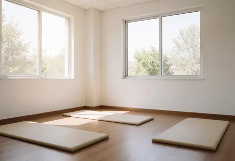 Intérieur d'une salle de soins vide équipée de tapis de sol sur un plancher en bois, éclairée par la lumière naturelle entrant par de grandes fenêtres et aux murs blancs.