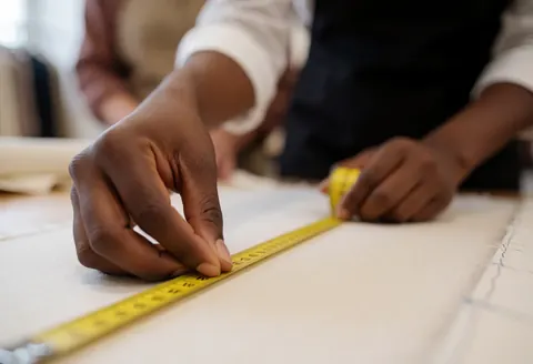 Une main utilise un mètre ruban jaune pour mesurer un tissu blanc dans un atelier de couture, avec un arrière-plan flou.
