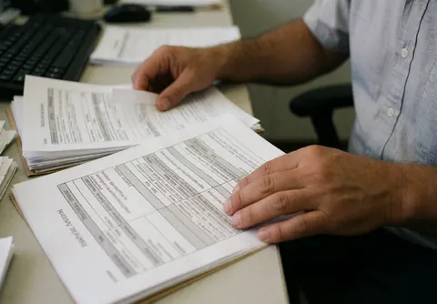 Un homme en chemise bleue consulte des papiers financiers sur un bureau équipé d'un clavier et d'une souris.