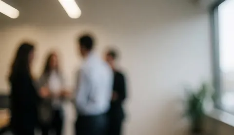 Groupe de professionnels discutant près d'une fenêtre dans un environnement de travail lumineux avec des plantes et un fond neutre.