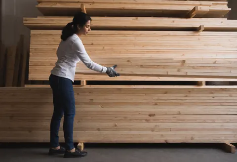 Une femme portant des gants organise des piles de bois dans un entrepôt de quincaillerie, baignée d'une lumière diffuse et dans une ambiance professionnelle.