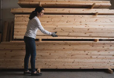 Une femme portant des gants organise des piles de bois dans un entrepôt de quincaillerie, baignée d'une lumière diffuse et dans une ambiance professionnelle.