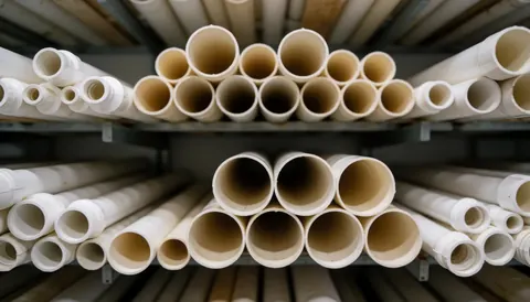 Des tubes blancs rangés sur des étagères métalliques dans un entrepôt industriel pour matériaux de construction.