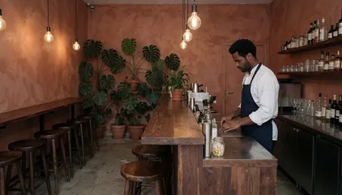 Intérieur d'un bar moderne avec un long comptoir en bois, des plantes en pot, des chaises et un serveur préparant des boissons.