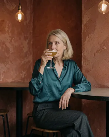 Une femme d'âge mûr profite d'un cocktail dans un bar aux murs de terre cuite et aux ampoules Edison, assise à une table avec une allure relaxée et élégante.