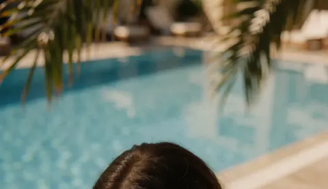 Une personne observe une piscine bleue avec des feuilles de palmier en premier plan dans un cadre relaxant et ensoleillé.