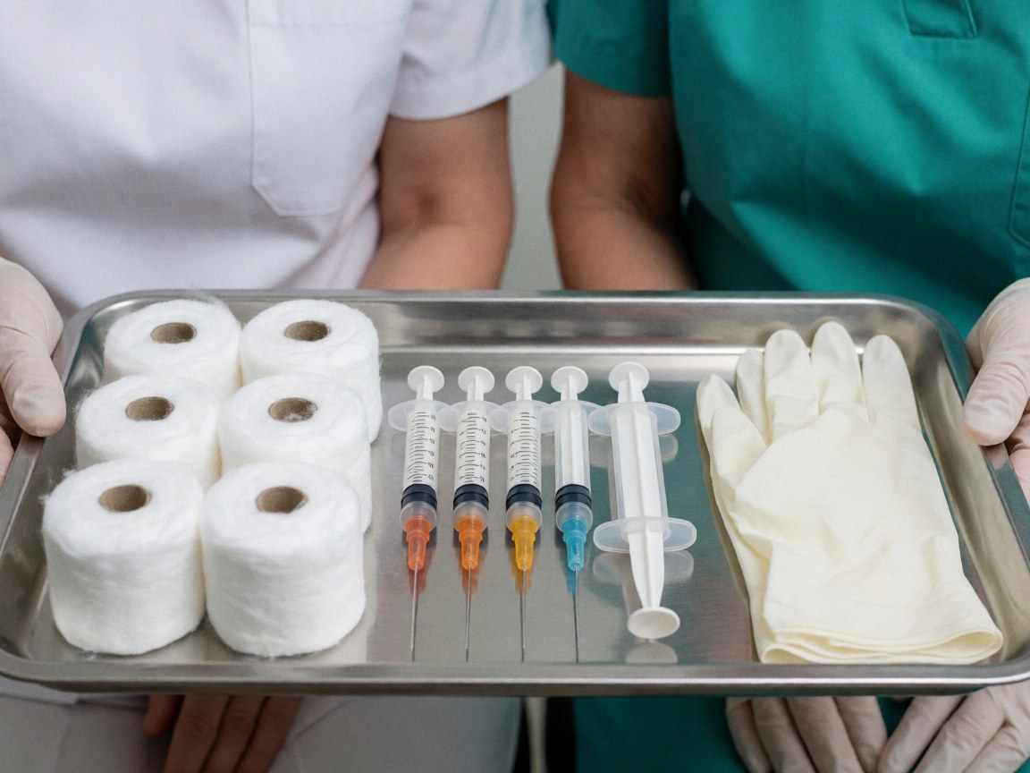 Deux professionnels de santé tiennent un plateau métallique contenant des seringues, des gants et du coton pour des interventions cliniques.