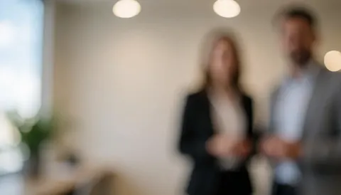 Deux professionnels en réunion dans un bureau moderne aux murs clairs et à l'éclairage doux, créant une ambiance corporative neutre.