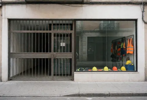 Façade d'un magasin de construction avec porte métallique et fenêtre montrant casques et gilets de sécurité. Murs blancs et trottoir propre.