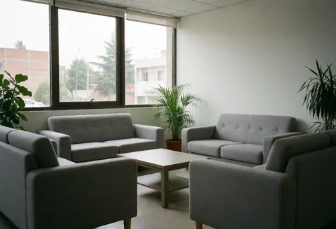 Intérieur d'une salle d'attente avec canapés gris, table basse, plantes vertes et grandes fenêtres laissant entrer la lumière naturelle.