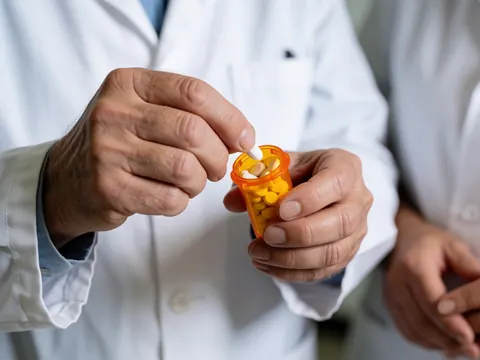 Un pharmacien prend des pilules d'un flacon orange avec les deux mains, une autre personne est visible en arrière-plan.