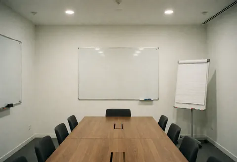 Salle de réunion moderne avec table en bois, chaises noires, tableaux blancs et murs blancs.