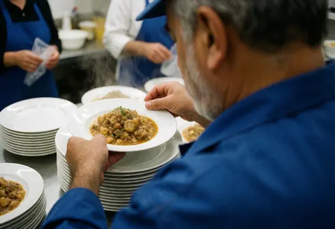 Un chef cuisinier mûr à la barbe sert un plat fumant dans une cuisine commerciale avec d'autres membres du personnel en arrière-plan.