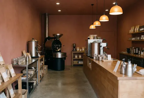 Vue intérieure d'une cafétéria montrant un comptoir en bois, un torréfacteur industriel, une cafetière et des produits de café disposés sur des étagères.