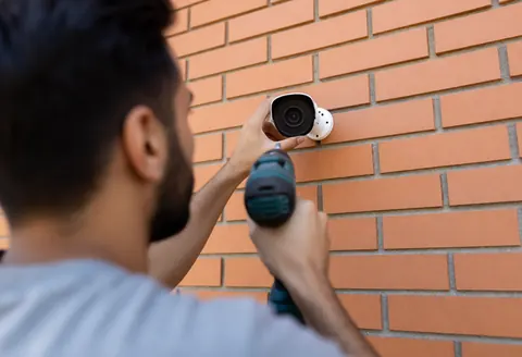 Un homme installe une caméra de surveillance sur un mur en briques à l'aide d'une perceuse électrique.