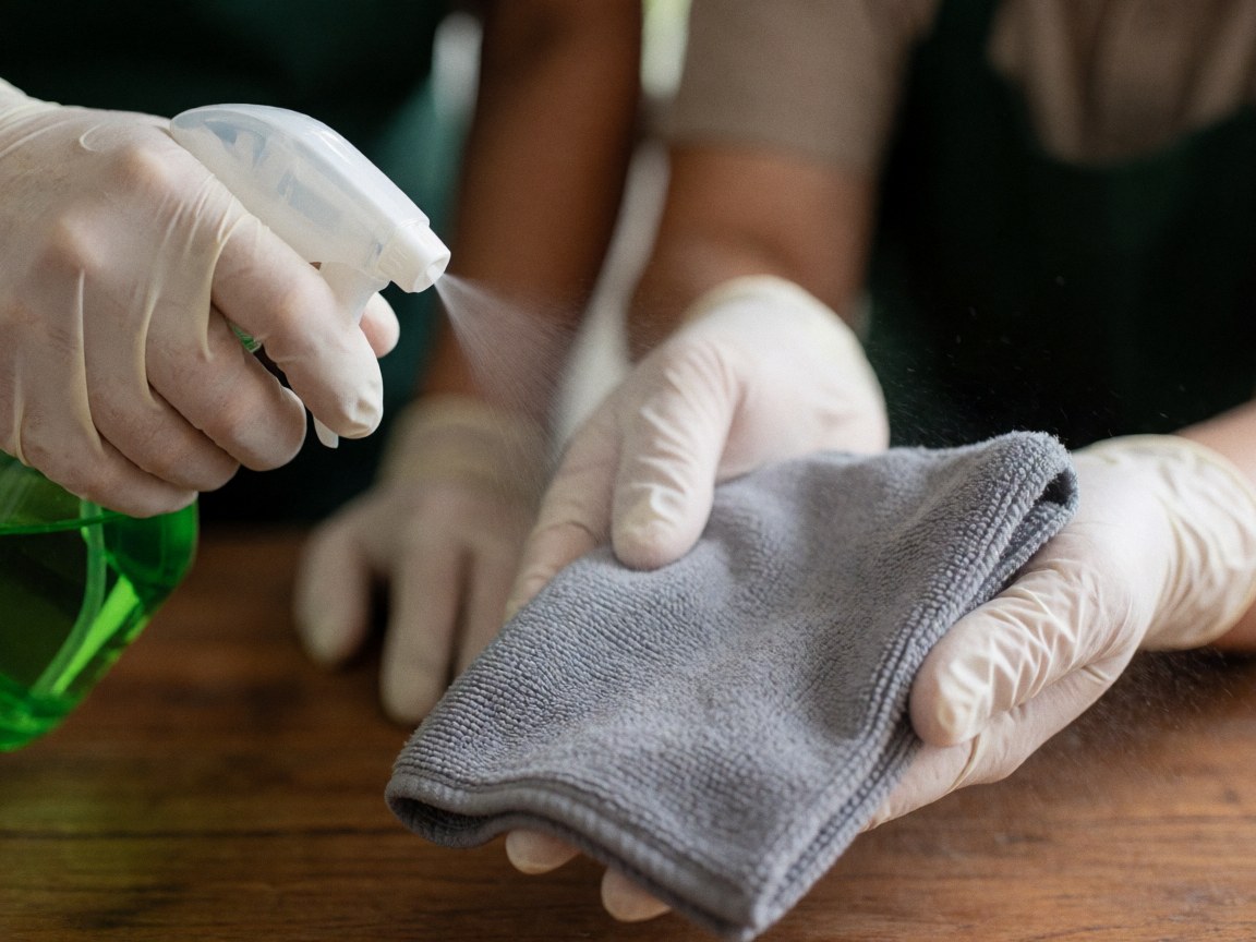 Une main portant un gant en latex pulvérise un liquide depuis un flacon sur un tissu gris tenu par l'autre main, posé sur une surface en bois.